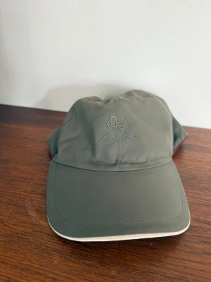 Olivgrön keps från Loro Piana - Snygg olivgrön keps från Loro Piana med broderad logga framtill. Kepsen har böjd skärm med ljus kant och är tillverkad i ett tunt, lätt material som känns sportigt och modernt. Perfekt accessoar för en avslappnad och stilren look.