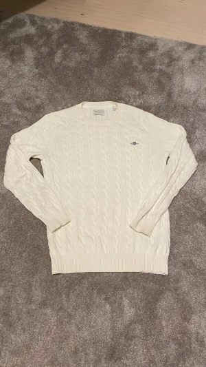 Vit kabelstickad tröja från Gant - Kabelstickad vit tröja från Gant med rund halsringning och broderad logotyp på bröstet. Tröjan har ribbade muddar vid ärmslut och nederkant. Klassisk och stilren design som passar till mycket. Materialet är mjukt och bekvämt.