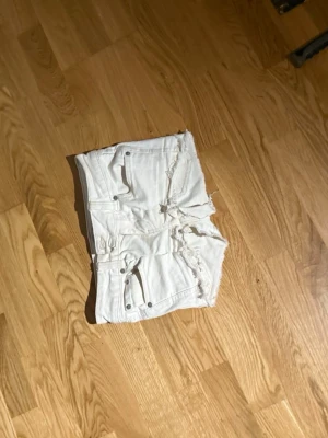 Vita jeansshorts med slitningar - Snygga vita jeansshorts med råa kanter och slitna detaljer. Klassisk femficksmodell med silverfärgade nitar och knapp. Perfekta för varma dagar och en avslappnad stil.