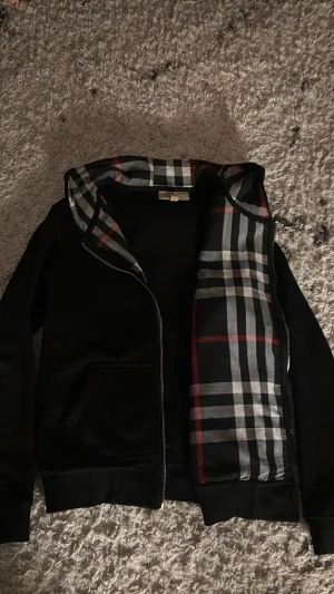 Svart hoodie från Burberry - Snygg svart hoodie från Burberry med dragkedja och klassiskt rutigt foder i huvan och insidan. Hoodien har två fickor framtill och ribbade muddar vid ärmar och nederkant. Perfekt för dig som gillar stilrena plagg med exklusiva detaljer.