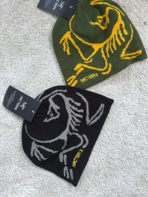 Arc'teryx mössa med logga - Två snygga mössor från Arc'teryx, en i olivgrönt med gul logga och en i svart med grå logga. Båda har stor grafisk logotyp och är tillverkade i mjukt stickat material. Perfekt för dig som gillar outdoor och streetwear.
