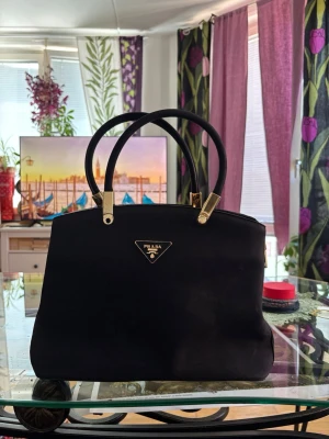 Svart handväska från Prada - Stilren svart handväska från Prada med klassisk trekantig logotyp i guld framtill. Väskan har två rundade handtag och guldfärgade metalldetaljer. Tillverkad i slätt läder som ger en lyxig känsla och passar perfekt till en trendig look.