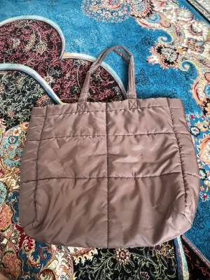 Brun quiltad shoppingväska - Säljer en brun quiltad shoppingväska med två långa handtag. Väskan har en mjuk, puffig känsla och rutmönstrad söm. Perfekt för dig som vill ha en rymlig och trendig tote till vardagen. Ingen synlig logga eller märke.