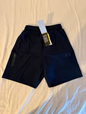 Under Armour träningsshorts – M passar S - Säljer ett par snygga och bekväma träningsshorts från Under Armour. Lätt och snabbtorkande material som passar perfekt för gym, löpning, padel eller annan träning. Resår i midjan med justerbar dragsko för optimal passform.  ✔️ Storlek: M men passar S ✔️ Skick: Helt nya med prislappar kvar ✔️ Färg: Svart ✔️ Lätt, ventilerande och bekvämt material  Hör gärna av dig om du har frågor eller vill ha fler bilder!