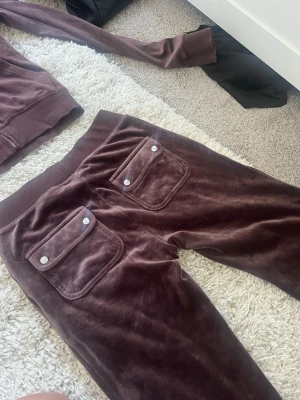 Bruna velourbyxor från Juicy Couture - Mörkbruna mjukisbyxor i mjuk velour från Juicy Couture. Byxorna har raka ben, bred resår i midjan och två bakfickor med lock och silverfärgade knappar. Perfekta för en chill och trendig look.