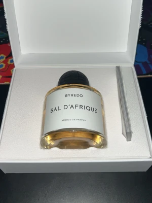 Byredo Bal d'Afrique Absolu 100ml och 102 ml mojave ghost samples - Byredo Bal d'Afrique Absolu de Parfum 100 ml i en elegant rund glasflaska med svart kork och vit etikett. Doften har en lyxig känsla och flaskan levereras i en stilren vit presentask. Perfekt för dig som gillar exklusiva parfymer med modern design.