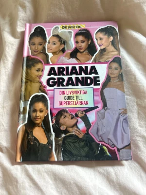 Ariana Grande: Din livsviktiga guide - En färgstark bok om superstjärnan Ariana Grande, fylld med bilder, fakta och roliga listor. Perfekt för fans som vill veta allt om hennes liv, musik, stil och karriär. Omslaget är rosa med collage av Ariana i olika outfits. Innehåller guider, topplistor och exklusiva insikter.
