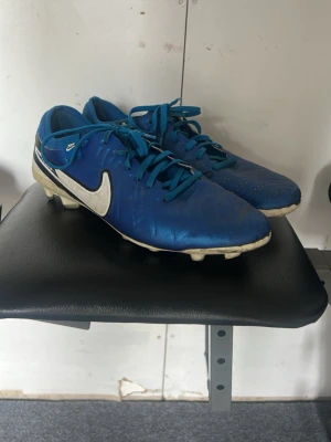 Blå Nike fotbollsskor med dobbar - Säljer ett par blå Nike fotbollsskor med vita detaljer och svart Nike-logga på sidan. Skorna har klassisk snörning och slitstarka dobbar för bra grepp på planen. Ovandelen är i syntetmaterial och sulan är vit med svarta detaljer. Perfekta för dig som vill ta spelet till nästa nivå.