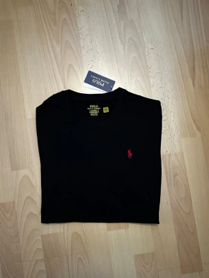 Svart t-shirt från Polo Ralph Lauren - Storlek M helt ny.Klassisk svart t-shirt från Polo Ralph Lauren med röd broderad logga på bröstet. Modellen är slim fit och har rund halsringning. Tillverkad i mjuk bomull för en skön känsla och stilren look.