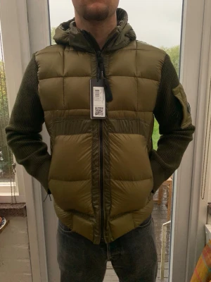 Olivgrön pufferjacka med huva - Snygg olivgrön pufferjacka med stickade ärmar och stor vadderad huva. Jackan har dragkedja framtill, ficka på ärmen och quiltad front. Perfekt för kalla dagar och ger en modern streetwear-look.