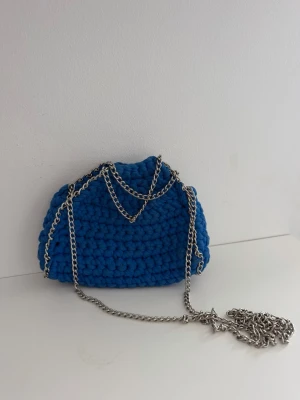 Blå virkad handväska med kedja - Cool blå handväska i virkat material med en chunky silverfärgad kedja som axelrem. Väskan har en mjuk, rundad form och ger en trendig och unik look. Perfekt för dig som vill sticka ut med accessoarer i färg. Aldrig använd, köpt från en tjej på plick som gör dem.