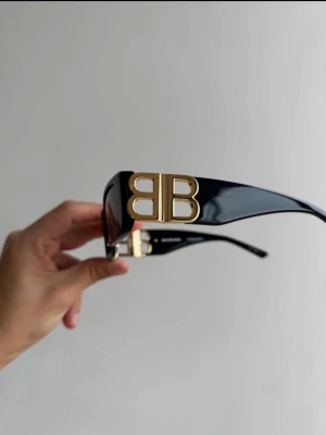 Svarta Balenciaga solglasögon  - Snygga svarta solglasögon från Balenciaga med rektangulära bågar och stora guldiga BB-loggor på sidorna. Glasögonen har en stilren och modern look med breda skalmar. Perfekta för dig som vill sticka ut med en lyxig accessoar.
