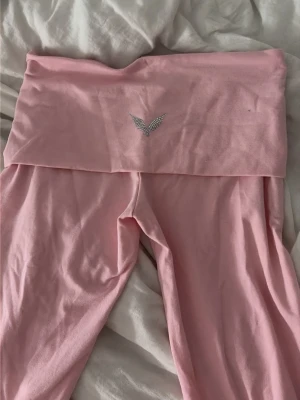 Victorias secret yoga pants  - Säljer ett par ljusrosa leggings med bred midjeresår och en snygg strassdetalj framtill. Mjukt och stretchigt material som sitter skönt på kroppen. Perfekta för en avslappnad och trendig look.