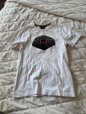 Vit t-shirt från Zavetti Canada - Snygg vit t-shirt från Zavetti Canada med ett stort broderat märke på bröstet och svart text samt röd lönnlöv-detalj. Klassisk rund halsringning och korta ärmar. Perfekt till jeans eller shorts för en clean och stilren look. Står att det är strlk S, men den är liten så skulle snarare säga att det är XS.