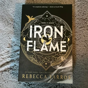 Iron Flame av Rebecca Yarros - Iron Flame är en fantasyroman med svart och guld omslag, dekorerad med drakmotiv och detaljerade mönster. Bokens sidor har svarta silhuetter av drakar längs kanten, vilket ger en unik och cool look. Perfekt för dig som gillar episka äventyr och snygga böcker i bokhyllan.