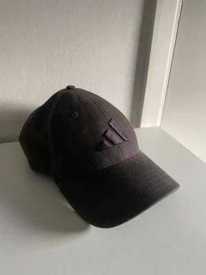 Svart keps från Adidas - Säljer en svart keps från Adidas med det klassiska broderade logotypen framtill. Kepsen har böjd skärm och justerbar rem bak. Tillverkad i bomullstyg som ger en avslappnad och sportig look.