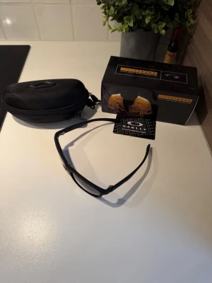 Svarta Oakley Livestrong solglasögon - Säljer ett par svarta Oakley Livestrong solglasögon med sportig design. Bågarna är mattsvarta och har Oakley-loggan på sidan. Linsen är mörktonad och glasögonen kommer med originalfodral och box. Perfekta för dig som vill ha en cool och funktionell stil.