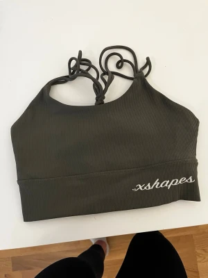 Olivgrön ribbad sport-bh xshapes - Säljer en olivgrön sport-bh från xshapes med ribbad struktur och tunna axelband som knyts i nacken. Bh:n har brottarrygg och vit broderad logga framtill. Perfekt för gymmet eller yoga.