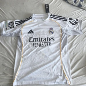 Real Madrid matchtröja Mbappé 10 - Säljer en vit Real Madrid matchtröja från Adidas med svarta detaljer och gula linjer. Tröjan har klubbmärke, Adidas-logga och Emirates Fly Better-tryck på framsidan. På ryggen står det Mbappé och nummer 10. Tillverkad i lätt och ventilerande material.