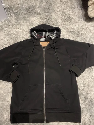 Burrbery  - Svart hoodie från Burberry med dragkedja framtill och klassiskt rutigt foder i huvan och längs insidan. Tröjan har två stora fickor fram, ribbade muddar och justerbar huva med snören. Perfekt för en chill och stilren look.