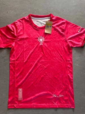 Röd x Rosa Portugal fotbollströja Nike - Snygg röd x rosa fotbollströja från Nike med Portugals landslagsemblem på bröstet och diskret logga på ärmen. Tillverkad i lätt och ventilerande polyester, perfekt för träning eller match. Kort ärm och klassisk passform. Märket 'The Skin' syns nedtill på framsidan.
