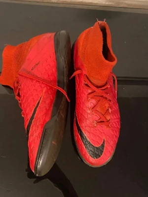 Nike HypervenomX röda fotbollsskor - Säljer ett par Nike HypervenomX fotbollsskor i rött med svart sula och svart Nike-logga. Skorna har en strumpliknande krage för extra stöd runt ankeln och snörning framtill. Ovandelen är i syntetmaterial med struktur för bättre bollkontroll. Perfekta för inomhusspel.