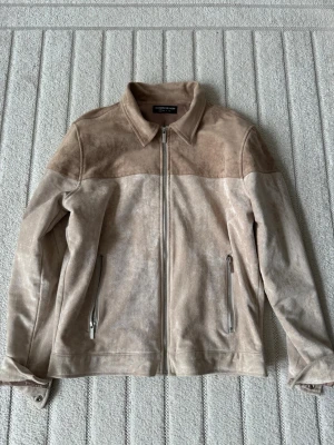 Beige skinnjacka från Manière De Voir - Snygg beige skinnjacka från Manière De Voir i mockaliknande skinn. Jackan har raka linjer, krage och två dragkedjeförsedda fickor framtill. Perfekt för dig som vill ha en stilren och modern look.