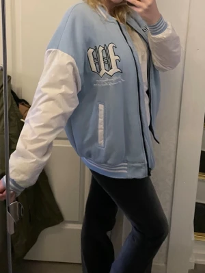 Ljusblå vårjacka med coola detaljer  - Snygg bomberjacka i ljusblått och vitt från Sports Studio. Jackan har vita ärmar, ribbade muddar och krage med blåvita ränder samt broderade detaljer på bröstet. Två fickor framtill och dragkedja. Perfekt för en sportig och trendig look. S. 