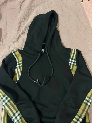 Burberry hoodie - Säljer nästan helt oanvänd Snygg svart hoodie med klassiska beige, röda och svarta rutiga detaljer längs ärmar och axlar. Huva med dragsko och mjukt material som känns skönt mot huden. Perfekt för dig som gillar streetwear och vill sticka ut lite extra.