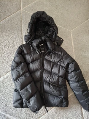 Svart quiltad pufferjacka från GANT - Snygg svart pufferjacka från GANT med huva och dragkedja framtill. Jackan har ett quiltat mönster och en patch med GANT-logga på ärmen. Perfekt för kalla dagar och riktigt skön att bära.