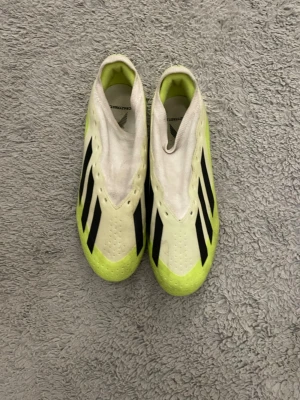 Adidas Crazyfast vita fotbollsskor - Säljer ett par Adidas Crazyfast fotbollsskor i vitt och neongult med svarta detaljer. Skorna har en strömlinjeformad design utan snörning och är tillverkade i syntetmaterial för bästa grepp och känsla på planen. Perfekta för dig som vill ha speed och kontroll.