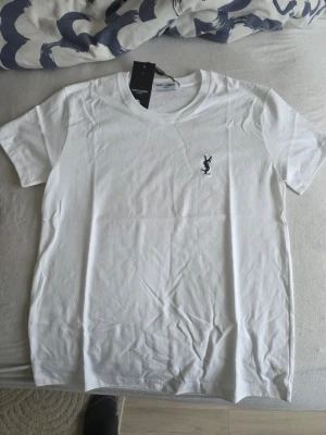 Vit t-shirt från Saint Laurent - Klassisk vit t-shirt från Saint Laurent med svart logga broderad på bröstet. T-shirten har rund halsringning och korta ärmar. Tillverkad i mjuk bomull för en skön känsla och clean look.