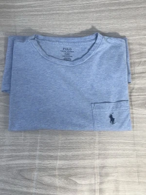 Ljusblå t-shirt från Polo Ralph Lauren - Snygg ljusblå t-shirt från Polo Ralph Lauren med klassisk rund halsringning och liten bröstficka med broderad logga. Tillverkad i mjuk bomull som känns skön mot huden. Perfekt till jeans eller shorts för en clean och enkel stil.