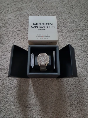 Omega x Swatch MoonSwatch Desert - Snygg Omega x Swatch MoonSwatch Desert från Bioceramic MoonSwatch Collection. Klockan har en beige boett, brun urtavla med vita detaljer och tachymeter, samt matchande beige armband med Omega-logga. Perfekt för dig som gillar unik design och ikoniska samarbeten.