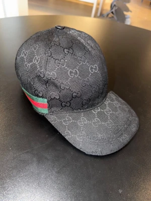 Svart Gucci keps med GG-mönster - Snygg svart keps från Gucci med det klassiska GG-monogrammet över hela kepsen. På sidan finns en grön och röd rand som ger en ikonisk touch. Kepsen har böjd skärm och är tillverkad i ett slitstarkt tygmaterial.