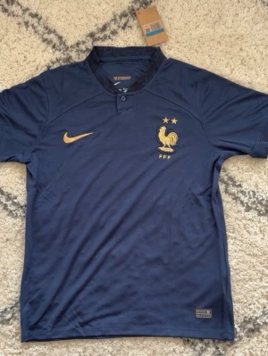 Frankrike Nike fotbollströja blå - Mörkblå fotbollströja från Nike med Frankrikes landslagsemblem och två stjärnor på bröstet. Guldiga detaljer och Nike-logga. Tillverkad i ett lätt och ventilerande material, perfekt för träning eller match.