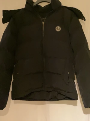 Trapstar Jacka Svart  - Säljer en äkta svart pufferjacka från Trapstar med hög krage och avtagbar huva. Jackan har dragkedja framtill, två sidofickor med knappar och Trapstar-logga på bröstet. Perfekt för kalla dagar och riktigt snygg! 