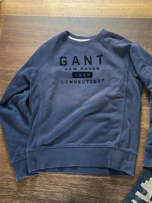 Blå sweatshirt från GANT - Mörkblå sweatshirt från GANT med tryck på bröstet där det står 'GANT New Haven 1949 Connecticut'. Tröjan har rund halsringning, långa ärmar och ribbade muddar vid ärmslut och nederkant. Perfekt för en avslappnad och stilren look.