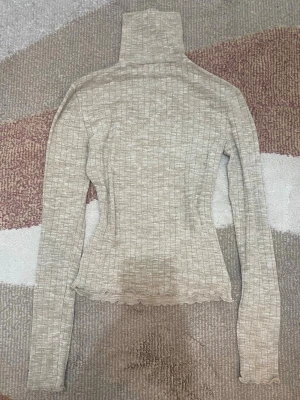 Beige ribbad polotröja - Säljer en beige ribbad stickad polotröja med lång ärm och figurnära passform. Tröjan har hög krage och vågig kant längst ner för en extra detalj. Perfekt att styla lager på lager under kalla dagar.