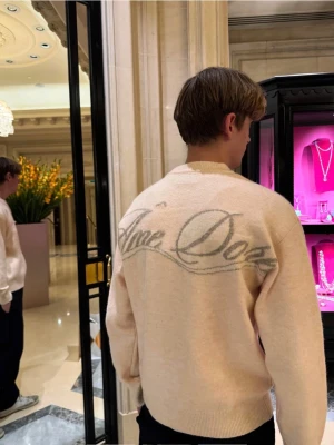 Beige stickad tröja från Ame Doré  - Stickad tröja i beige från Ame Doré med rund halsringning och ribbade muddar. Tröjan har en stor logga i grått på ryggen och en mindre logga framtill. Mjuk och stilren design som passar till det mesta.