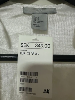 Vit spetskofta från H&M - Säljer en elegant vit kofta från H&M med vackert spetsmönster och lång ärm. Koftan är öppen framtill och har en lätt glansig innerdel som ger en lyxig känsla. Perfekt för att lyfta en outfit med en romantisk touch.