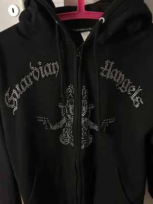 Svart hoodie med strass och tryck - Säljer en svart hoodie från BLINGLUCID med dragkedja, huva och snörning. Framsidan har texten 'Guardian Angels' och ett motiv i strass som ger en riktigt edgy vibe. Perfekt för dig som gillar streetwear och detaljer som sticker ut. Pris kan diskuteras ( säljs inte längre) 