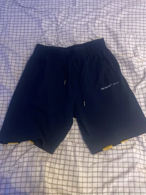 Off-White svarta shorts med tryck - Säljer ett par mörkblå shorts från Off-White med vita och gula pilar på baksidan och diskret logga framtill. Shortsen har resårmidja med snörning och är perfekta för sommaren. Materialet känns mjukt och bekvämt. Bomull är materialet på demhär shortsen