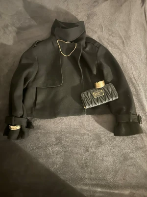 Svart kort ullrock med detaljer - Stilren svart kort rock med hög krage och axelklaffar. Rocken har snygga spännen vid ärmsluten och en asymmetrisk knäppning framtill. Tillverkad i ett tjockare ullmaterial som ger en exklusiv känsla och passar perfekt till höst och vinter. Går att bära på tre olika sätt. Pris går att diskutera!