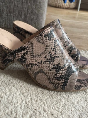Slip-in skor med ormmönster - Säljer ett par slip-in klackar från Graceland med snyggt ormmönster i beige och svart. Skorna har öppen tå och bred klack, tillverkade i konstläder med struktur som liknar ormskinn. Perfekta för dig som vill sticka ut med ett trendigt mönster. Storlek 40 men mer 39.