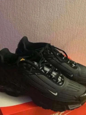 Nike Tuned 1 svarta sneakers - Svarta Nike Tuned 1 sneakers med diskreta vita detaljer och klassisk swoosh på sidan. Skorna har en sportig siluett, robust sula och snörning framtill. Materialet är en mix av syntet och textil för en modern look.