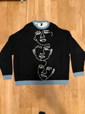 Svart stickad tröja med ansikten - Cool svart stickad tröja med tre vita abstrakta ansikten broderade på framsidan. Tröjan har ljusblå ribbade muddar vid ärmslut, nederkant och halsringning. Perfekt för dig som gillar unik design och streetstyle.