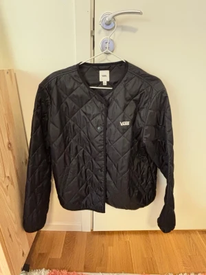 Svart quiltad vindjacka från Vans - Snygg svart vindjacka från Vans med quiltat mönster och rund halsringning. Jackan har tryckknappar framtill och en diskret Vans-logga på bröstet. Tillverkad i lätt, glansigt material som ger en sportig look. Perfekt för lager-på-lager-stil. Passar stl S-L beror på hur man vill att den ska sitta.
