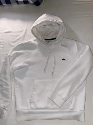 Vit hoodie från Lacoste med logga - Snygg vit hoodie från Lacoste med den klassiska gröna krokodilloggan broderad på bröstet. Hoodien har dragsko i huvan och en stor magficka framtill. Tillverkad i mjukt bomullsmaterial och har ribbade muddar vid ärmslut och nederkant.