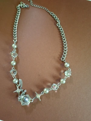Silverfärgat halsband med pärlor och stjärnor - Snyggt halsband i silverfärg med kedja, dekorerat med vita pärlor, klara stenar och stjärnformade detaljer. Perfekt accessoar för att lyfta din outfit med en touch av havsinspirerad stil. Materialet är metall och plastpärlor.